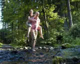 Ein Mann trägt eine Frau auf dem Rücken durch einen Bach im Wald. Die Sonne scheint durch die Bäume. Ein idyllischer Ort, wie eine teuto quelle.