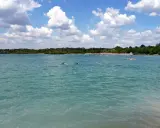 Schwimmer im türkisfarbenen Wasser des Beckum Sees, mit bewaldetem Ufer und Wolken am Himmel.