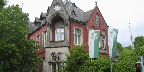 Die Kaiservilla in Minden, ein rotes Backsteingebäude mit Türmchen und zwei GWD-Fahnen, umgeben von üppigem Grün.