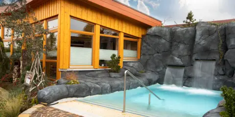Entspannendes Wasserbecken mit Wasserfällen vor einer Holzhütte, ideal für eine H2O Sauna.