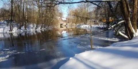 Winterliche Idylle an der **Planwiese Telgte**: Eisiger Fluss, schneebedeckte Ufer und ein Fachwerkhaus spiegeln sich im Wasser.
