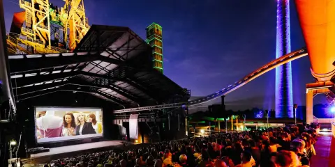 Ein belebtes **freiluft kino** mit vielen Zuschauern, die einen Film auf einer großen Leinwand vor einer beeindruckenden Industriekulisse mit bunten Türmen und Stahlkonstruktionen genießen.