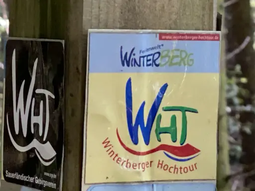 Schilder für die Winterberger Hochtour. Ein Schild zeigt "WHT" in Weiß auf Schwarz, das andere "Winterberg" und "Winterberger Hochtour" in bunten Farben.
