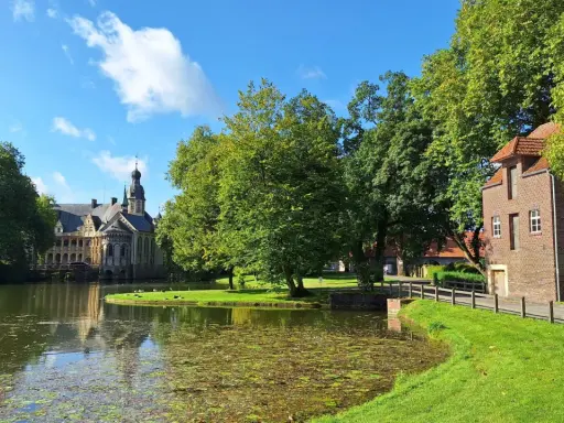 Ein idyllischer vechte radweg führt vorbei an einem Teich, grünen Bäumen und einem historischen Wasserschloss.