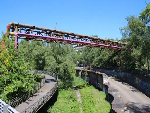 Alte Rohrleitungen der Erzbahntrasse Bochum ragen über üppiges Grün. Ein geschwungener Fußweg schlängelt sich durch die Natur.