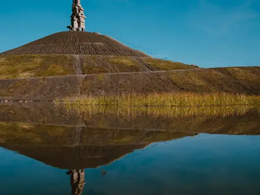 Spiegelung einer Halde im Ruhrgebiet mit einer Steinskulptur auf dem Gipfel und Schilf am Ufer.