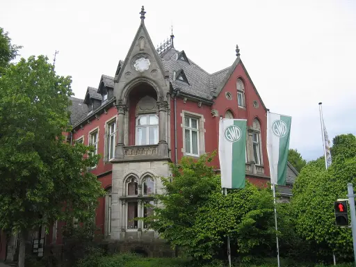 Die Kaiservilla in Minden, ein rotes Backsteingebäude mit Türmchen und zwei GWD-Fahnen, umgeben von üppigem Grün.