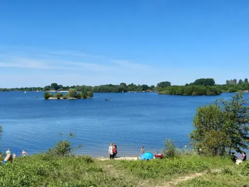 Sonniger Tag am Lippesee Paderborn. Menschen entspannen am Ufer, Segelboote gleiten über das Wasser, eine kleine Insel ziert die Mitte.