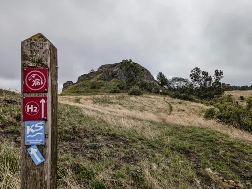Wanderwegweiser mit Falken-Symbol und "H2"-Markierung, der zu den **rundwanderwege owl** führt. Ein Pfad schlängelt sich über einen grasbewachsenen Hügel zu einer Felsformation.