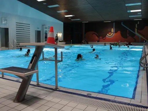 Menschen schwimmen im **Gartenhallenbad Enger**. Ein Sprungbrett mit einem orange-weißen Kegel steht am Beckenrand.