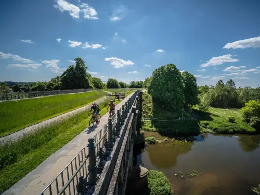 Zwei Radfahrer genießen die Aussicht auf der hohen Mark Radroute über eine Brücke, umgeben von grünen Wiesen und Bäumen.