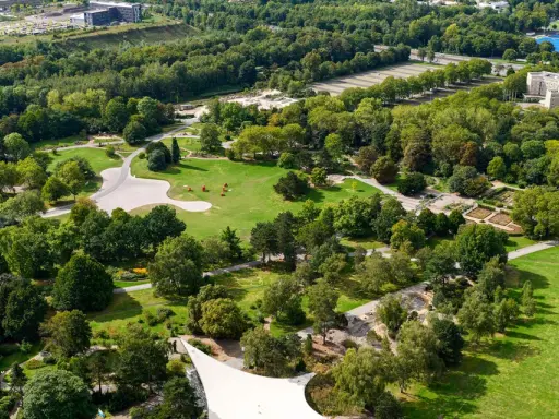 Luftaufnahme eines weitläufigen **Parks in Dortmund** mit grünen Rasenflächen, verschlungenen Wegen, blühenden Beeten und dichtem Baumbestand.