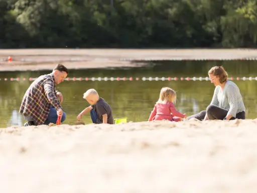 Familie spielt am Sandstrand des badesee mindenerwald. Vater hilft Sohn beim Sandburgen bauen, während Mutter und Tochter daneben sitzen.