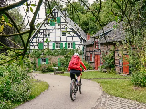 Radfahrerin auf einem Weg zwischen Fachwerkhäusern in NRW. Grüne Fensterläden und üppige Vegetation schmücken die idyllische Szenerie.