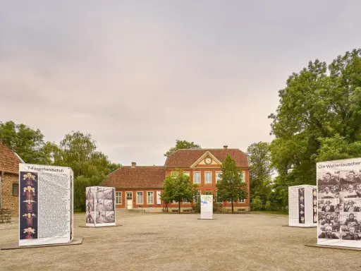 Das Haus Nottbeck mit Ausstellungen im Freien. Mehrere Schilder mit Fotos und Texten stehen vor dem roten Backsteingebäude.