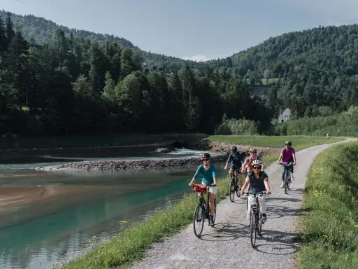 Gruppe Radfahrer auf einem Weg entlang eines Flusses mit bewaldeten Hügeln im Hintergrund.