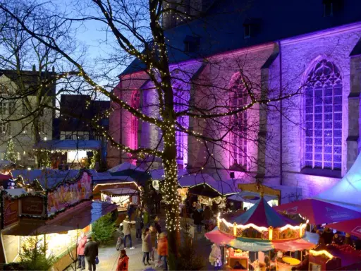 Bunte Lichter und Buden auf dem weihnachtsmarkt werne. Eine Kirche im Hintergrund erstrahlt in lila Licht.