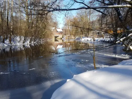 Winterliche Idylle an der **Planwiese Telgte**: Eisiger Fluss, schneebedeckte Ufer und ein Fachwerkhaus spiegeln sich im Wasser.