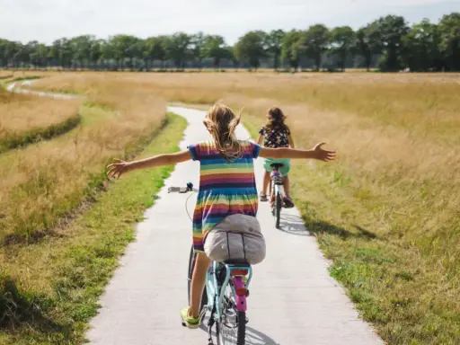 Zwei Mädchen genießen ein sonniges Fahrrad-Wochenende auf einem Feldweg, die Arme weit ausgebreitet.