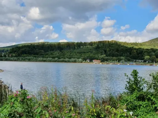 Idyllische Landschaft am Emmer-Radweg mit See, bewaldeten Hügeln und einigen Anglern am Ufer.