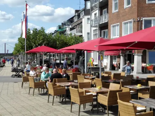 Gemütliche Straßencafés mit roten Sonnenschirmen laden zum Verweilen ein. Menschen spazieren am Rhein entlang, im Hintergrund die Brücke von Emmerich.