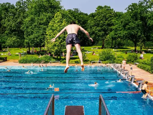 Ein Mann springt in ein Freibad in NRW. Viele Menschen sonnen sich auf der grünen Liegewiese im Hintergrund.