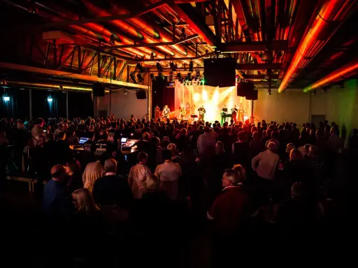 Ein Konzert in Emsdetten mit einer Band auf der Bühne und vielen begeisterten Zuschauern. Die Beleuchtung taucht die Szene in ein warmes, rotes Licht.