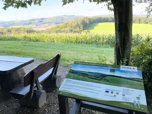Holzbänke und ein Tisch laden zum Verweilen ein. Ein Schild "Familienplatz - Weg der Blicke" weist auf die Aussicht hin.