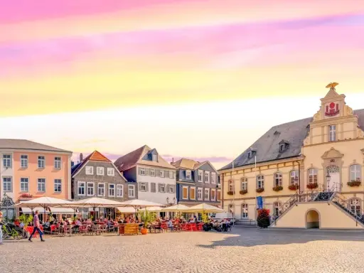 Ein Marktplatz mit Cafés und historischen Gebäuden, der an das "Venedig Westfalens" erinnert, unter einem farbenfrohen Himmel.