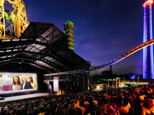 Ein belebtes **freiluft kino** mit vielen Zuschauern, die einen Film auf einer großen Leinwand vor einer beeindruckenden Industriekulisse mit bunten Türmen und Stahlkonstruktionen genießen.