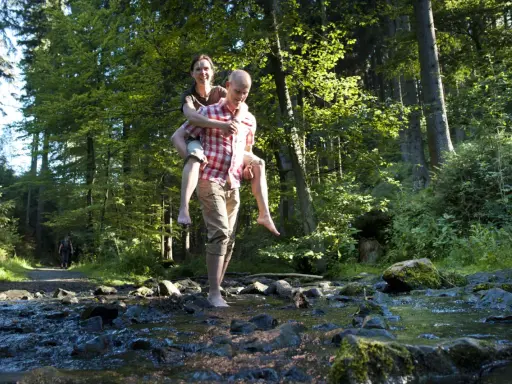 Ein Mann trägt eine Frau auf dem Rücken durch einen Bach im Wald. Die Sonne scheint durch die Bäume. Ein idyllischer Ort, wie eine teuto quelle.