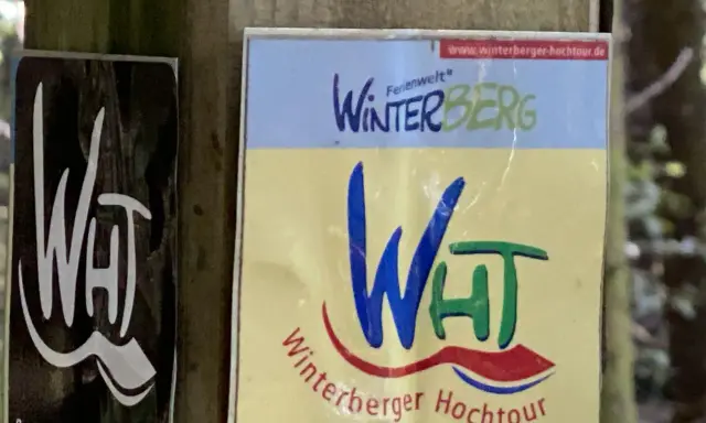 Schilder für die Winterberger Hochtour. Ein Schild zeigt "WHT" in Weiß auf Schwarz, das andere "Winterberg" und "Winterberger Hochtour" in bunten Farben.