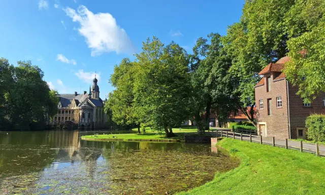Ein idyllischer vechte radweg führt vorbei an einem Teich, grünen Bäumen und einem historischen Wasserschloss.