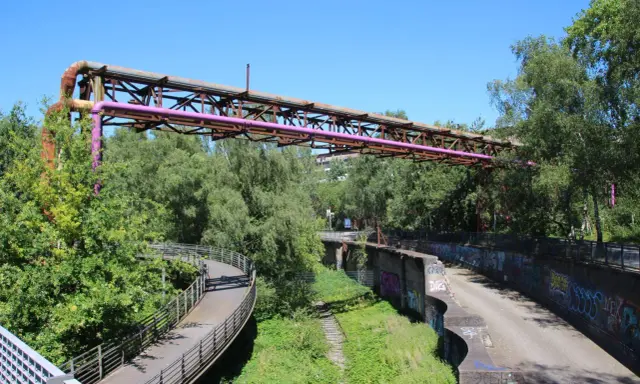Alte Rohrleitungen der Erzbahntrasse Bochum ragen über üppiges Grün. Ein geschwungener Fußweg schlängelt sich durch die Natur.