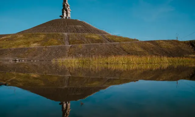 Spiegelung einer Halde im Ruhrgebiet mit einer Steinskulptur auf dem Gipfel und Schilf am Ufer.