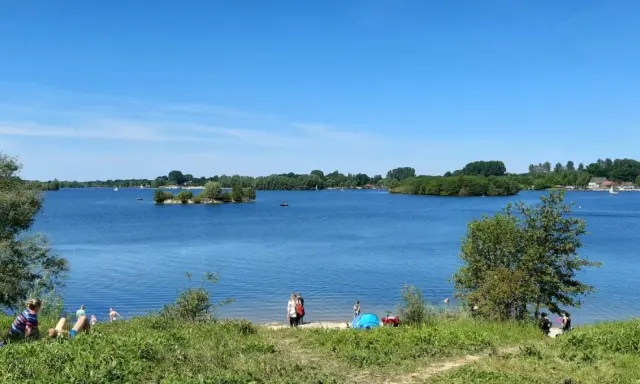 Sonniger Tag am Lippesee Paderborn. Menschen entspannen am Ufer, Segelboote gleiten über das Wasser, eine kleine Insel ziert die Mitte.