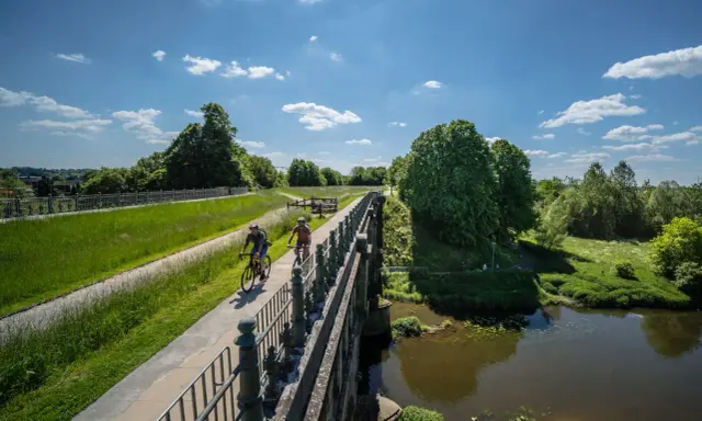 Zwei Radfahrer genießen die Aussicht auf der hohen Mark Radroute über eine Brücke, umgeben von grünen Wiesen und Bäumen.