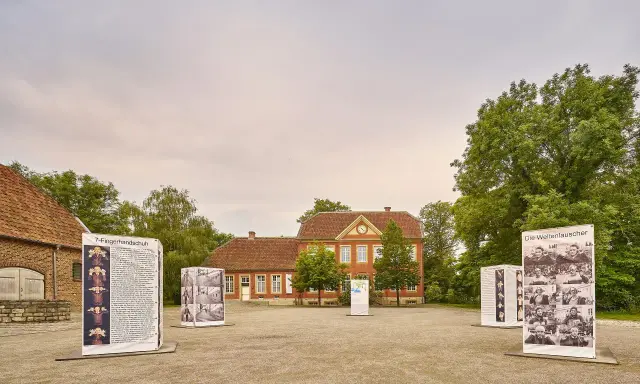 Das Haus Nottbeck mit Ausstellungen im Freien. Mehrere Schilder mit Fotos und Texten stehen vor dem roten Backsteingebäude.