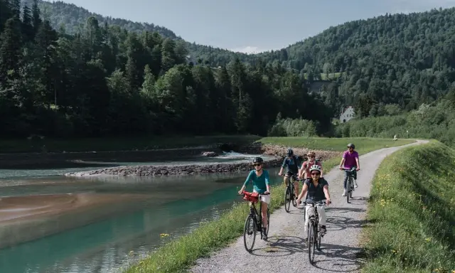 Gruppe Radfahrer auf einem Weg entlang eines Flusses mit bewaldeten Hügeln im Hintergrund.