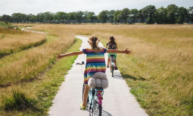 Zwei Mädchen genießen ein sonniges Fahrrad-Wochenende auf einem Feldweg, die Arme weit ausgebreitet.