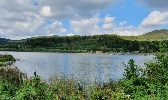 Idyllische Landschaft am Emmer-Radweg mit See, bewaldeten Hügeln und einigen Anglern am Ufer.