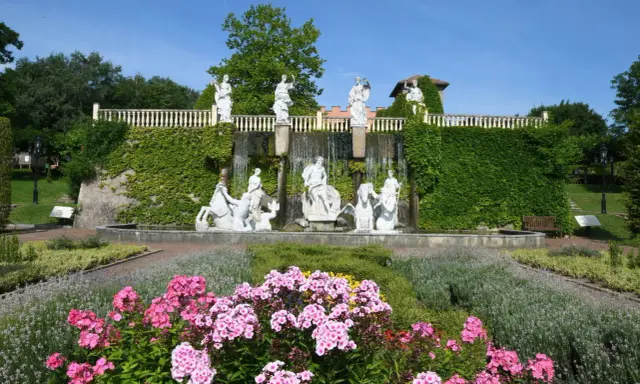 Prachtvoller Brunnen mit Skulpturen und Wasserfällen, umgeben von blühenden Blumen und Grün im botanischen Garten NRW.