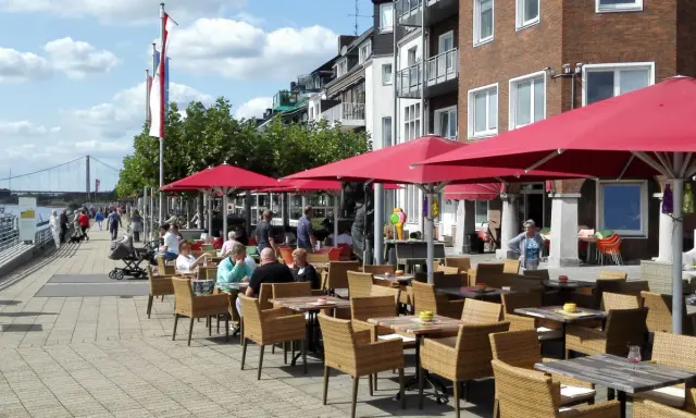 Gemütliche Straßencafés mit roten Sonnenschirmen laden zum Verweilen ein. Menschen spazieren am Rhein entlang, im Hintergrund die Brücke von Emmerich.