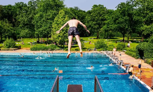 Ein Mann springt in ein Freibad in NRW. Viele Menschen sonnen sich auf der grünen Liegewiese im Hintergrund.