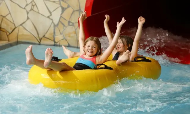 Zwei lachende Kinder rutschen auf einem gelben Schlauchboot durch einen Wasserpark. Ein perfekter Tag, wie ein h2o gutschein!