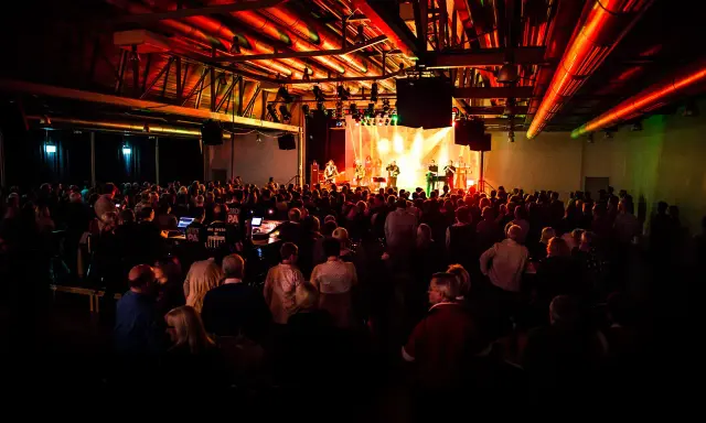 Ein Konzert in Emsdetten mit einer Band auf der Bühne und vielen begeisterten Zuschauern. Die Beleuchtung taucht die Szene in ein warmes, rotes Licht.