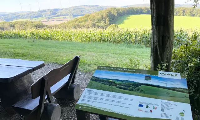 Holzbänke und ein Tisch laden zum Verweilen ein. Ein Schild "Familienplatz - Weg der Blicke" weist auf die Aussicht hin.