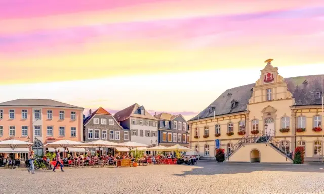 Ein Marktplatz mit Cafés und historischen Gebäuden, der an das "Venedig Westfalens" erinnert, unter einem farbenfrohen Himmel.