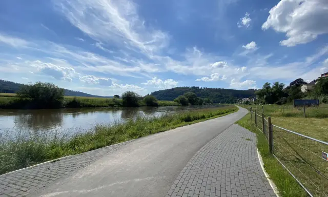 Ein asphaltierter Weg säumt den Fluss, ideal für eine Weser-Radweg Rundtour. Grüne Ufer und ein blauer Himmel mit Wolken laden zum Radfahren ein.