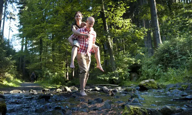 Ein Mann trägt eine Frau auf dem Rücken durch einen Bach im Wald. Die Sonne scheint durch die Bäume. Ein idyllischer Ort, wie eine teuto quelle.